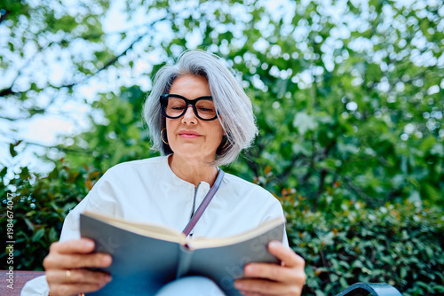 Schilderij op canvas Mature woman with grey hair and glasses quietly reading a book outdoors, indulgi