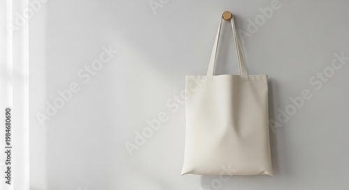 A plain white tote bag hangs on a hook against a light gray wall, casting a subtle shadow from natural light