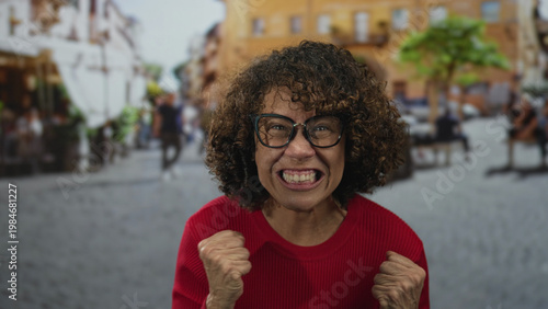 Obraz na plátně Woman wearing red sweater clenches fists amid outdoor cafe seating on city street under warm sunlight; excitement