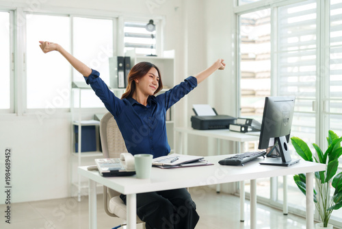Adult Asian business woman stretches arms upward taking work break feeling refreshed modern office workspace professional remote freelance worker wellbeing healthy posture at desk