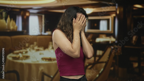 Bilde på lerret Young hispanic woman anxiously covers face while seated at polished wooden table in elegant restaurant dining area; stress