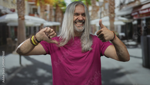 Fényképezés Man with long gray hair and magenta shirt shows thumbs up and thumbs down on street; ambivalence choice decision