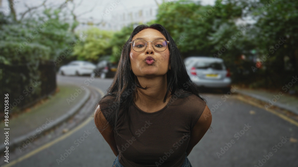 Fototapeta premium Young brunette woman wearing glasses points both index fingers toward camera showing hands and wrists on a leafy residential street; playful confidence.