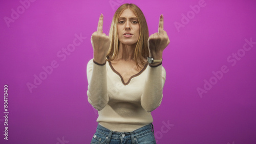 Obraz na plátně Woman raises middle fingers and flips off toward camera in studio with magenta backdrop, wearing cream sweater and denim jeans; anger defiance