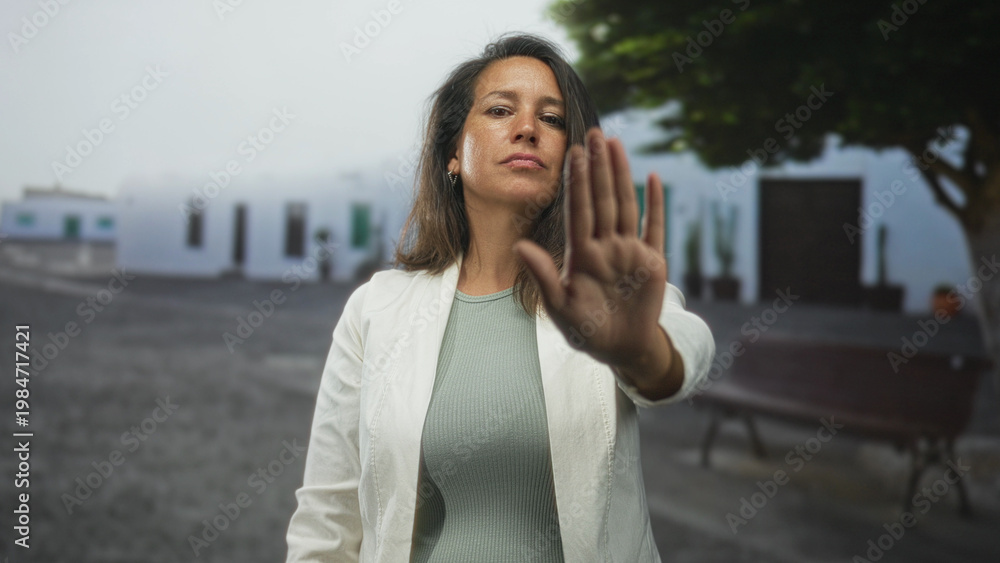 Fototapeta premium Woman holds up hand palm forward for stop in street in front of white building and bench; defiance protection.