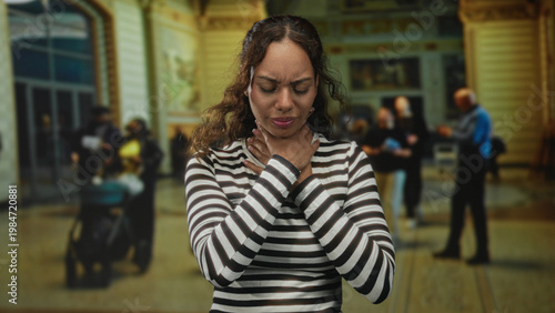 Bild auf Leinwand Young african american woman in a striped top holding her throat and grimacing in an ornate building concourse; choking emergency distress