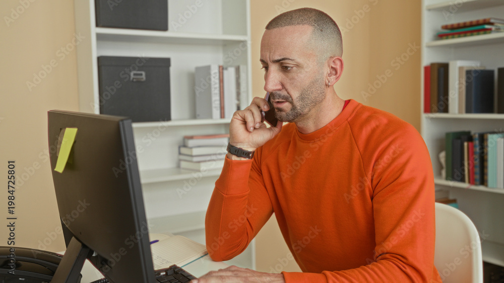 Fototapeta premium Man in orange sweater holds smartphone to ear and types on a computer keyboard at a desk with sticky note on monitor in a building office; remote work concern.
