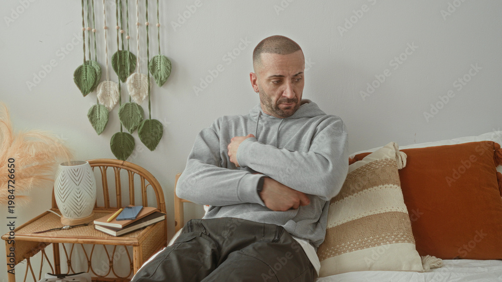 Fototapeta premium Man holds tablet to chest on bed in bedroom, arms crossed and looking down beside rattan side table with books and decorative pillow; quiet contemplation.