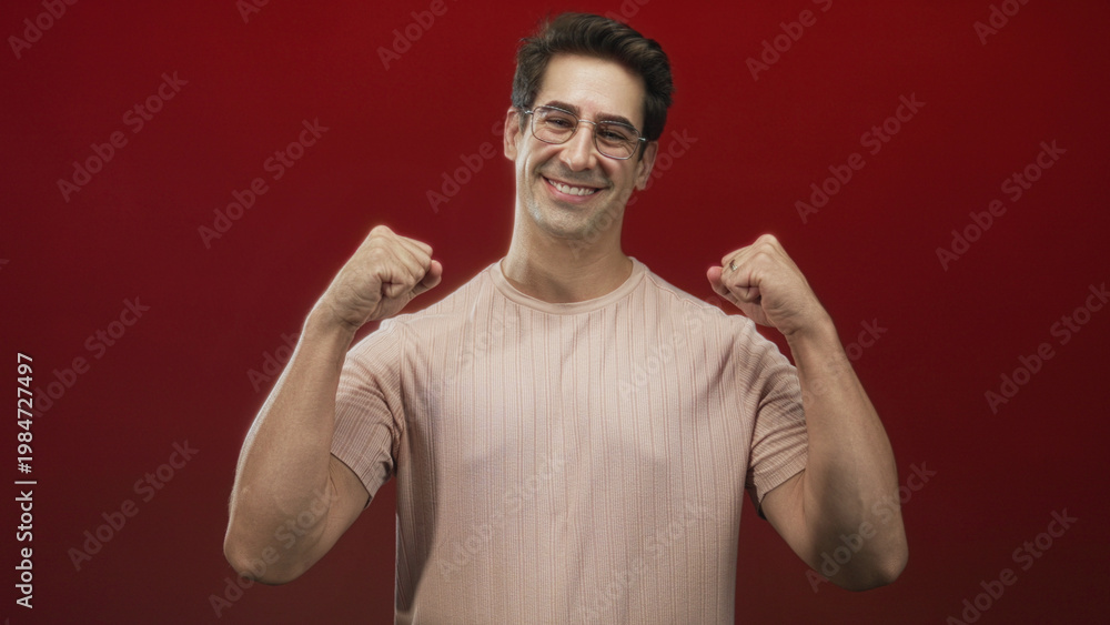 Naklejka premium Man raising clenched fists and smiling in a red studio wearing glasses and a casual t shirt; joy triumph.