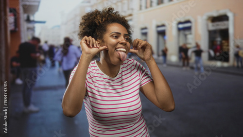 Fotografie Young african american woman in a striped t shirt pulls her cheeks and sticks out her tongue while making playful faces on a crowded city street; playful expression