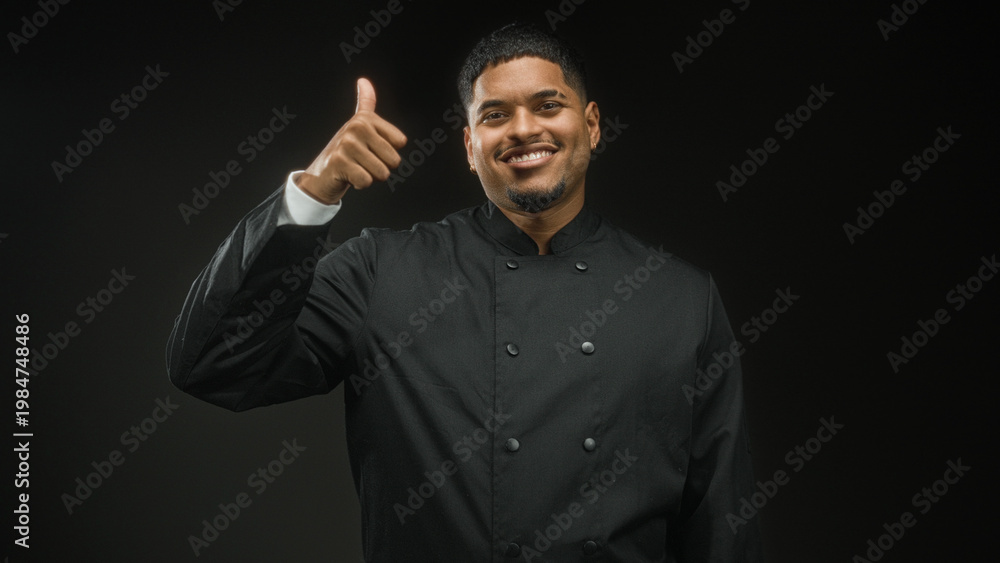 Obraz premium Hispanic man chef in black uniform showing peace sign and later thumbs up with hand in studio, smiling and wearing earring; confidence.