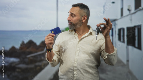 Fotografie Man holds creditcard in left hand and car key in right hand by a building on the seaside walkway; indecision choices