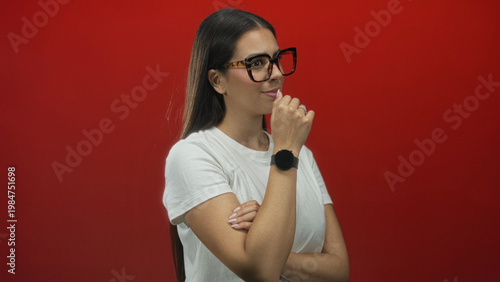 Εκτύπωση καμβά Woman in white t shirt with hand covering mouth and tortoiseshell glasses, arms crossed in red studio; pensive reflection