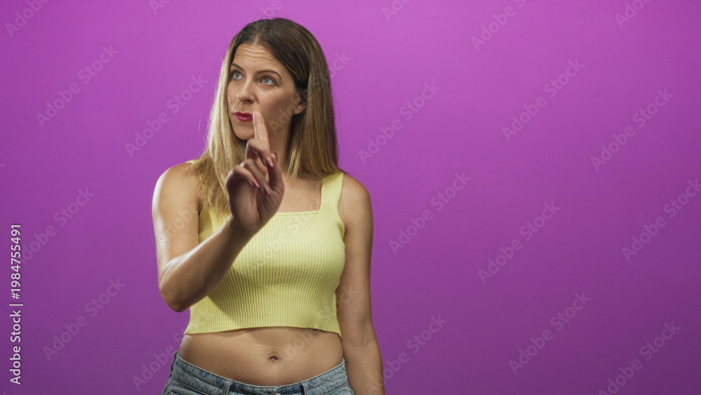 Obraz premium Woman points finger upward with exposed midriff wearing yellow crop top in studio smiling slightly; curiosity.