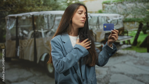 Young woman holds creditcard in right hand, left hand on chest showing thankful gesture, looking upward near parked golf cart on street; relief financial security.