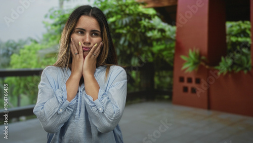 Woman pressing her cheeks with both hands on a building balcony, wearing a light blue blouse and yellow nails visible; thoughtful solitude.