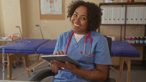 Quadro su tela Young african american woman doctor in blue scrubs sits in clinic building holding tablet with both hands wearing a red stethoscope around neck; compassion