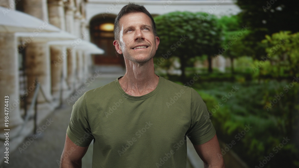 Obraz premium Man smiling and looking up beside a colonnade building with green park foliage and stone walkway, wearing olive tee; serenity health.