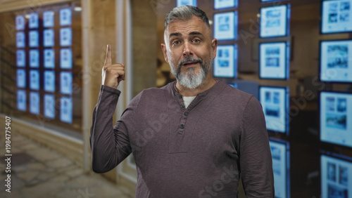 Obraz na plátně Man pointing finger to property display in building window while smiling and gesturing at real estate listings inside showroom; confidence sales pitch