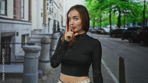 Quadro su tela Young woman with red hair in a black crop top showing bare midriff, finger to lips for silence on a city street; secret playfulness