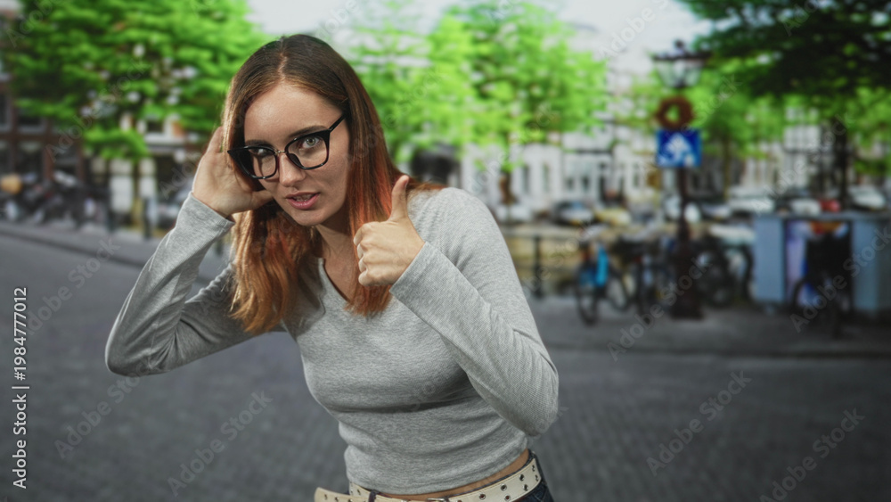 Fototapeta premium Young redhead woman with hand to ear giving thumbs up on a city street with bicycles and buildings visible; playful approval.