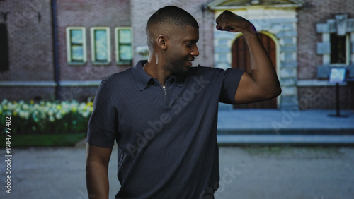 Tableau sur toile Man flexes bicep with arm bent at elbow, smiling and looking at his raised arm beside a stone building entrance with steps and tulip flowerbed; confidence strength