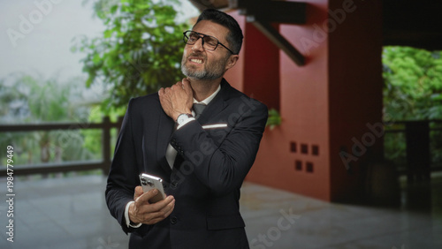 Fotografi Man in suit holding smartphone and rubbing neck in hotel building while checking messages; discomfort workday