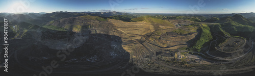 Aerial view of the massive open-pit mine terraces and winding industrial roads surrounded by green mountains in Santa Lucia de Gordon, Castile and Leon, Spain.