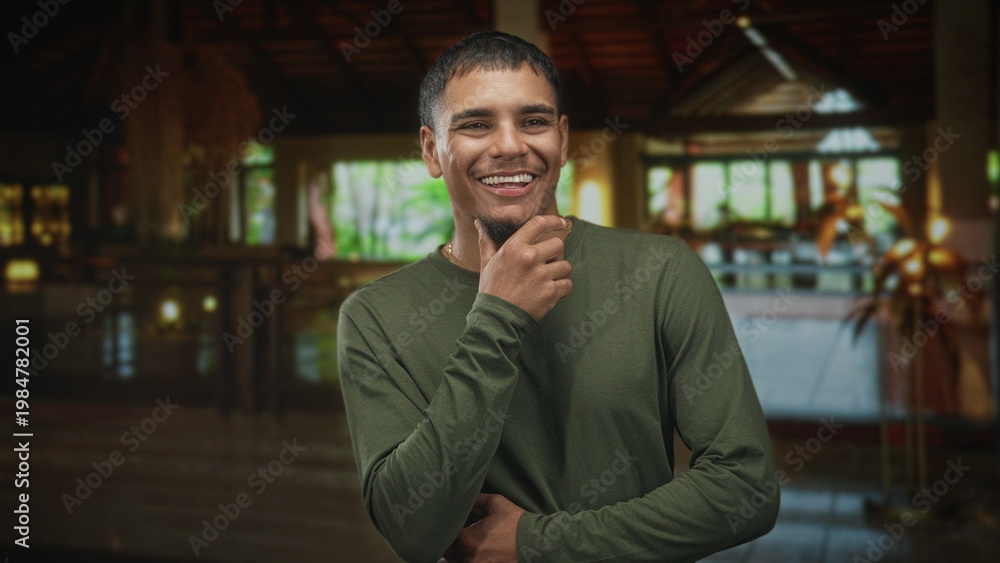 Naklejka premium Man touching mouth with his hand while smiling subtly in a building lobby; thoughtful quiet introspection.