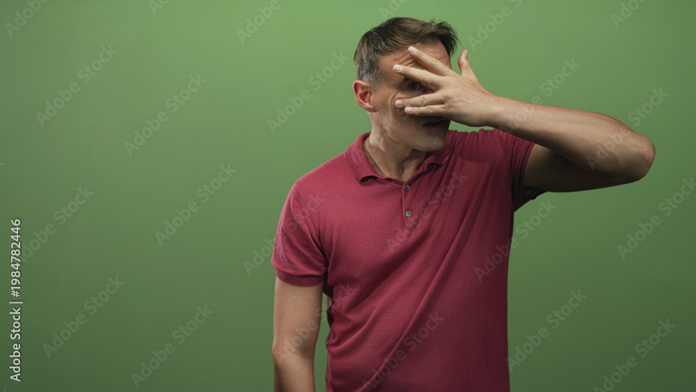 Fototapeta premium Man covering face with hand and peeking between fingers, wearing red polo against green screen in studio; embarrassment privacy vulnerability.