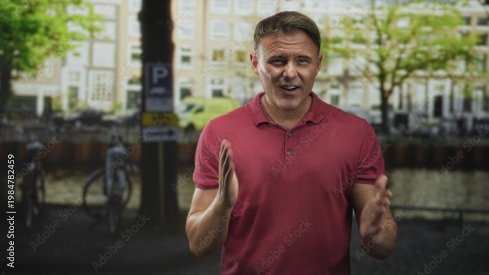 Fototapeta premium Man clapping hands beside parked bicycles on a canal street in front of brick townhouses, red polo visible; engaging presentation.