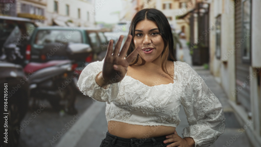 Fototapeta premium Woman showing four fingers with outstretched hand and slight smile, wearing white embroidered crop top and nose ring while standing on a narrow city street; playful confidence.