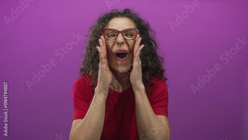 Obraz na plátně Young hispanic woman wearing red shirt with hands on cheeks and mouth agape against purple studio wall; surprise