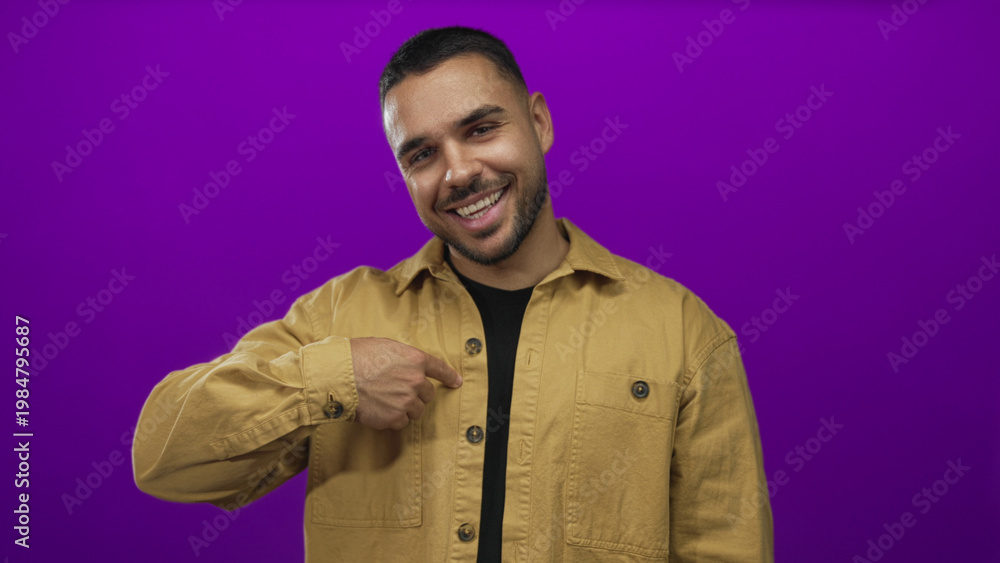Obraz premium Man wearing tan jacket smiles and points finger to shirt while giving thumbs up in purple studio; confidence.