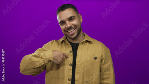 Fotografie Man wearing tan jacket smiles and points finger to shirt while giving thumbs up in purple studio; confidence