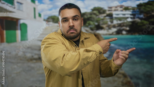 Man points both index fingers toward turquoise water beside a white building on sandy shore under bright sky; doubt.