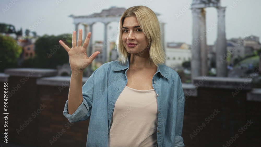Fototapeta premium Woman holds up hand showing palm and fingers while slightly smiling in front of stone columns on a building terrace, denim shirt visible; friendly greeting.