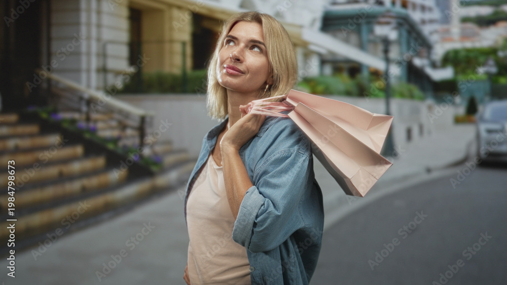 Fototapeta premium Woman young blonde holding pink shopping bags over shoulder, hand on hip, smiling and looking at camera while standing on street near steps; confidence leisure.