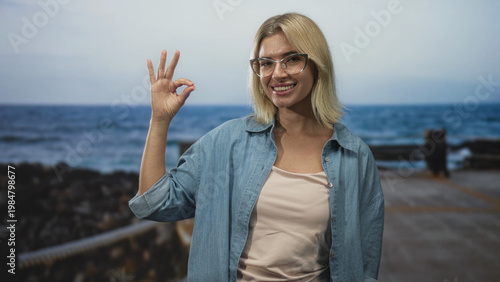 Fotografi Woman smiling and holding hand in ok sign in a studio with pier backdrop and denim shirt visible; confidence approval
