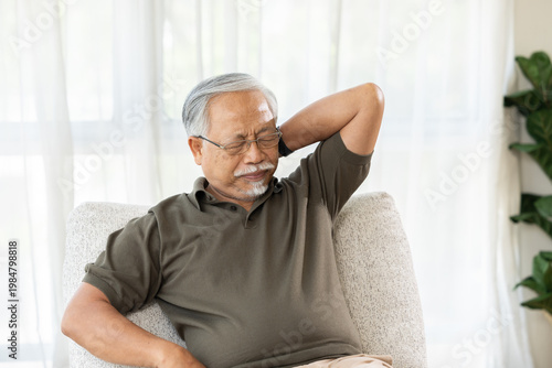 Senior Asian man experiencing severe neck pain and muscle ache while sitting on comfortable sofa at home feeling extremely unhappy and uncomfortable holding hand to stiff body