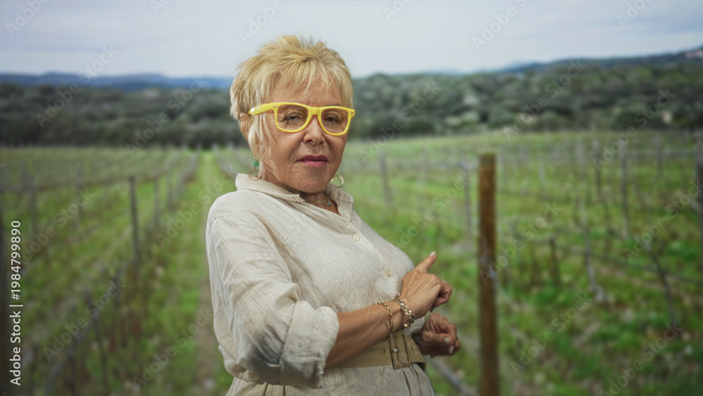Naklejka premium Woman points finger and checks wrist in a vineyard wearing yellow glasses and linen dress; thoughtful reflection.