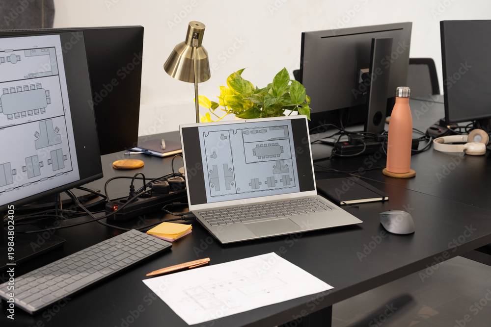 Fototapeta premium Silver laptop displaying floor plan on black desk in office with monitors, coral water bottle
