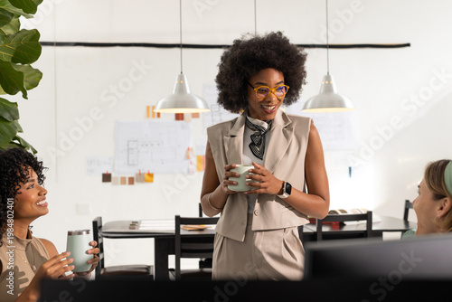 Diverse women team in office listening as standing woman in suit raising mug, discussing plans