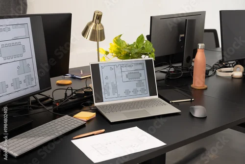 Fototapeta Silver laptop displaying floor plan on black desk in office with monitors, coral water bottle