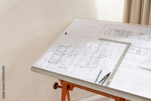 Drafting table holding plans, T-square, compass, pencil on alum edge with orange sawhorse in studio