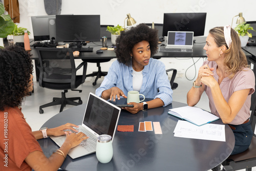 Diverse female coworkers discussing floor plans and swatches at round office table using tablet