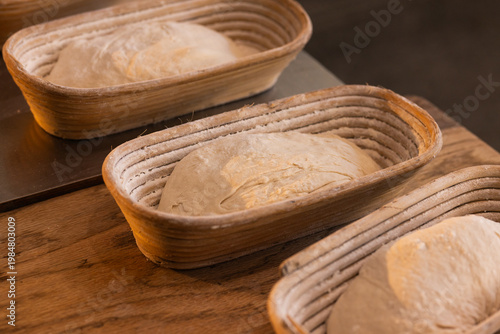 Oval woven bannetons holding three flour-dusted loaves of proofing dough on wooden countertop