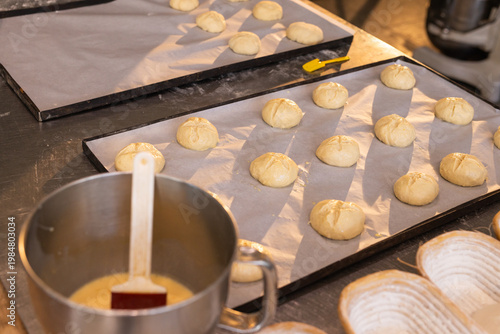 Two parchment-lined trays holding scored rolls are proofing on stainless counter with pastry brush