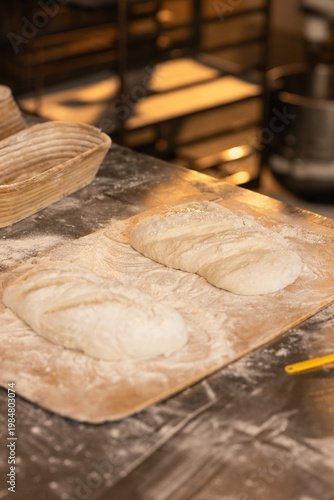 Two scored wheat loaves proofing on floured wooden peel at bakery prep table with yellow scraper