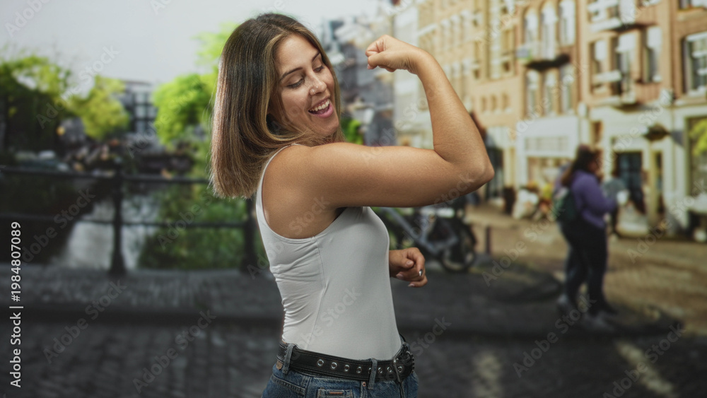 Fototapeta premium Woman flexes bicep and smiles in white tank top beside canal on a cobbled street with buildings and bicycle; confidence empowerment.